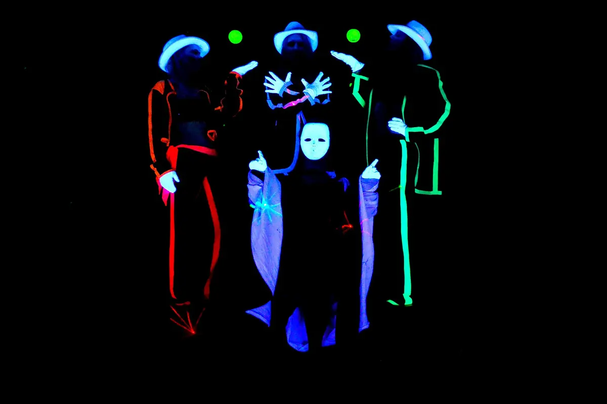 Four performers in hats and masks outlined in glowing red, blue and green stand against a black background in a Prague black light show