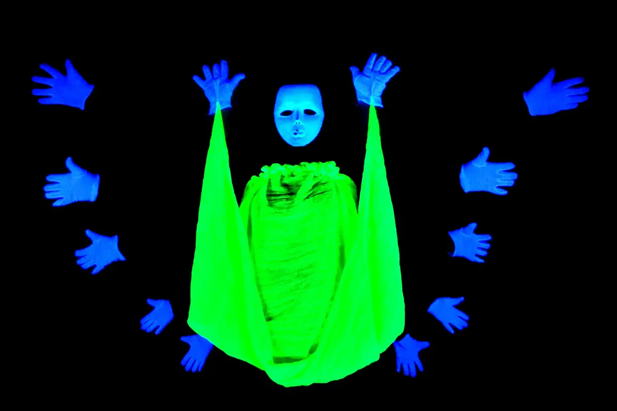 A glowing green cloth with a white mask on top, ringed by floating blue gloved hands, against complete blackness