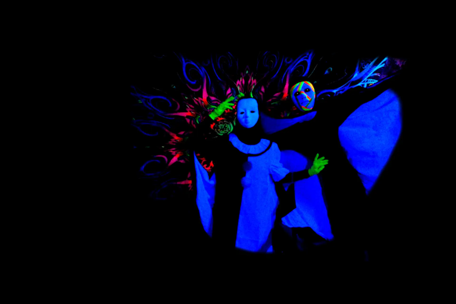 Two masked performers in glowing blue costumes and green gloves stand in front of a colourful fluorescent backdrop in a dark black light theatre show in Prague