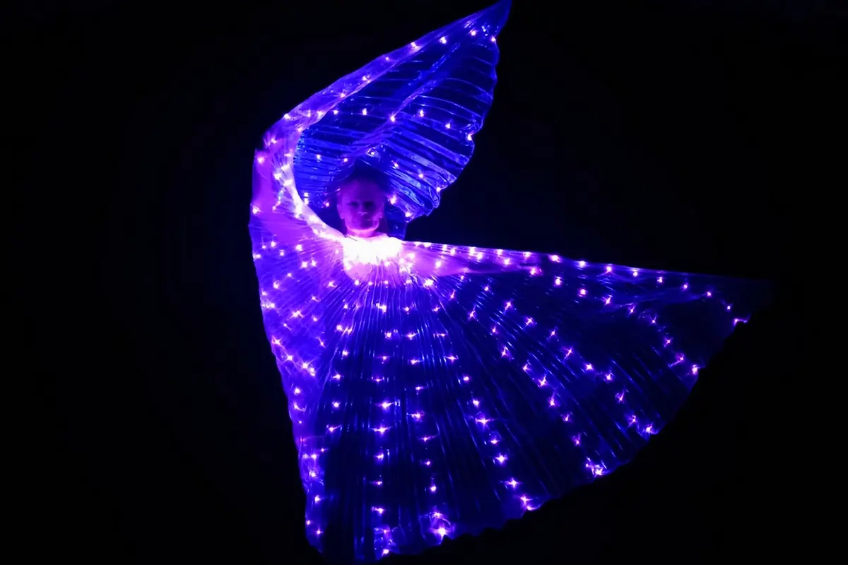 A performer in a fan of purple lights spreads giant illuminated wings in a dark theatre